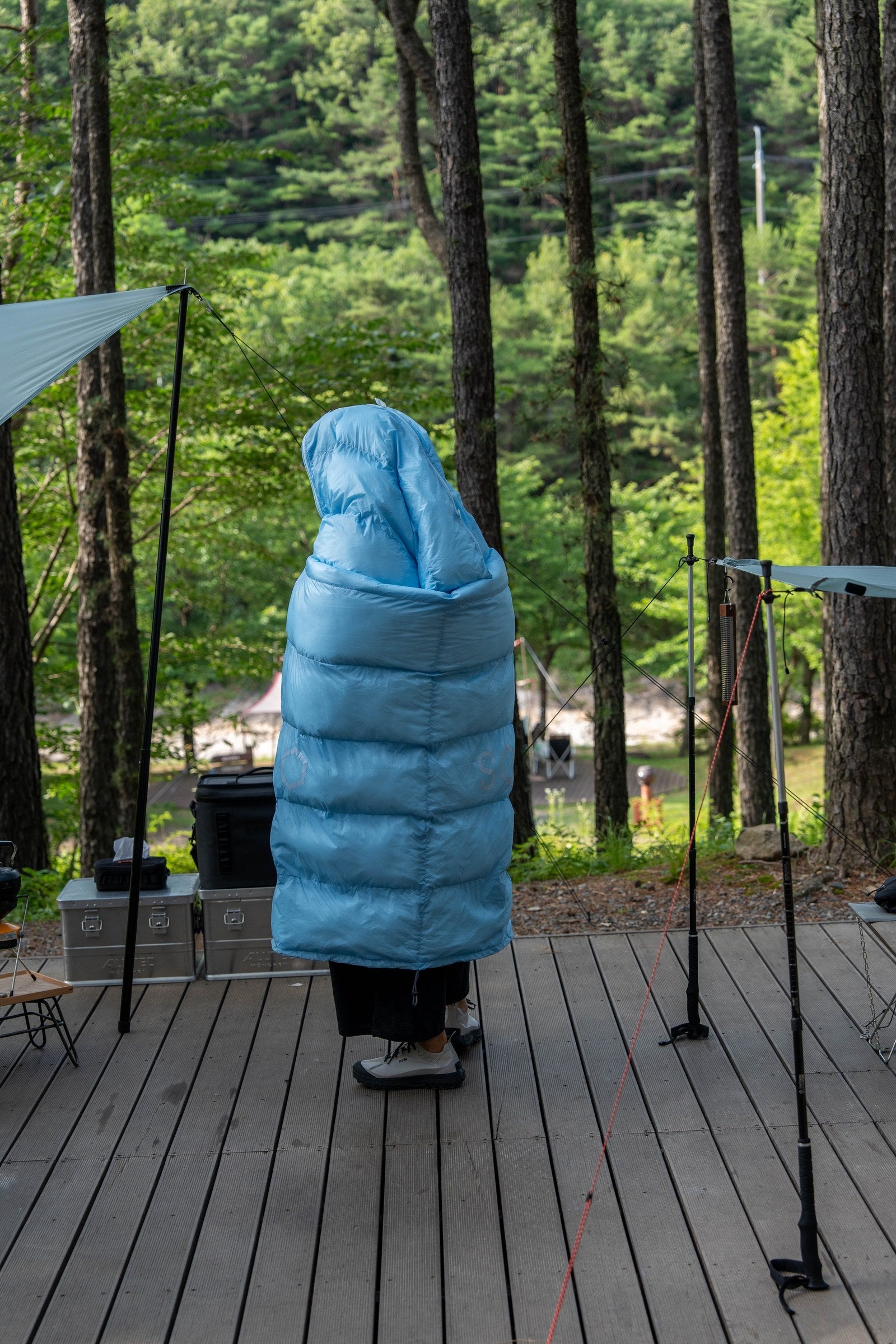 Camper wrapped in a sky blue DEOS camp comforter on a forest deck
