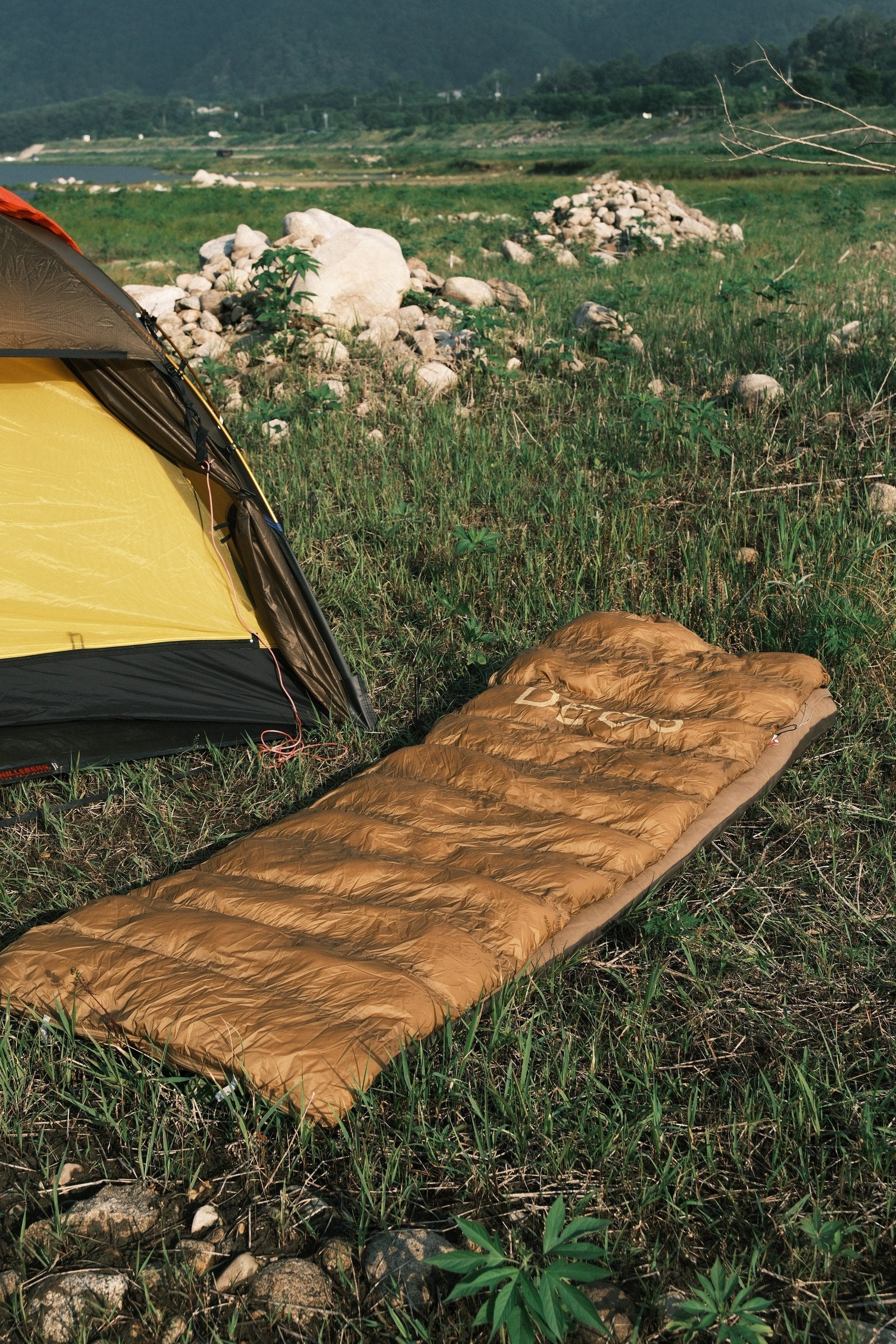 Tan DEOS camp comforter spread in a tent by the river