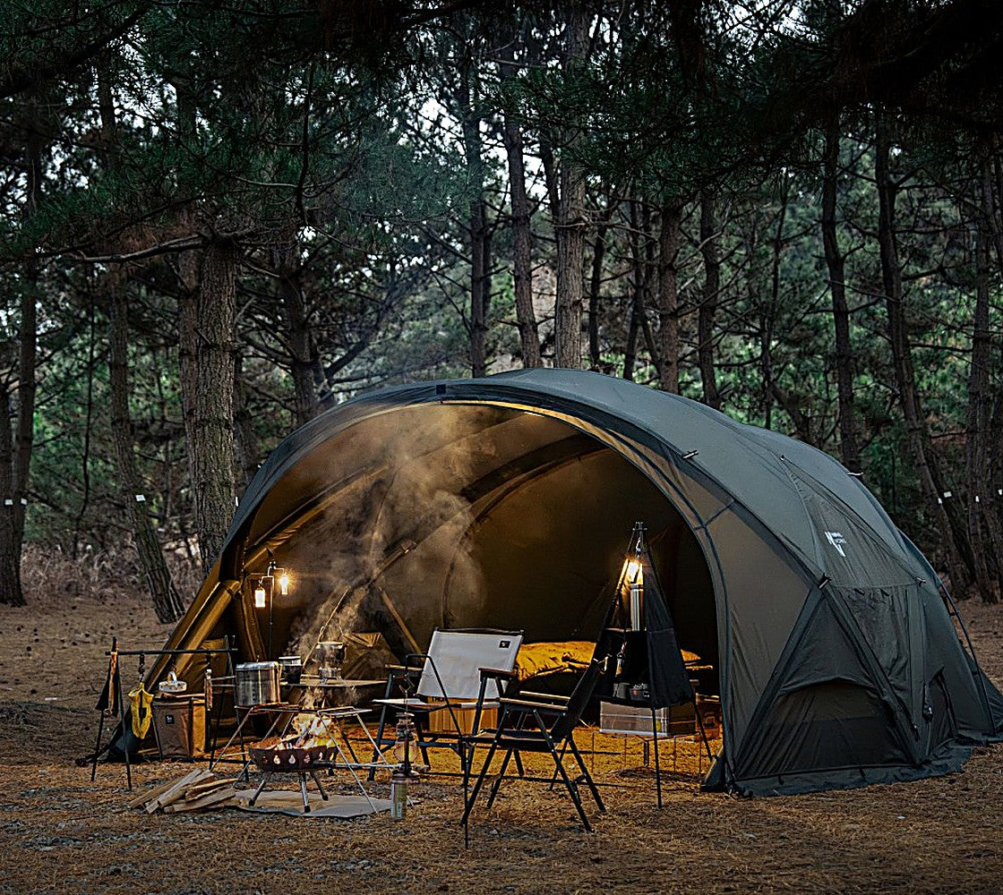 Evening camp with the Shelter GM tent's TPU Door open, lantern-lit bedding inside and campfire smoke in a pine forest