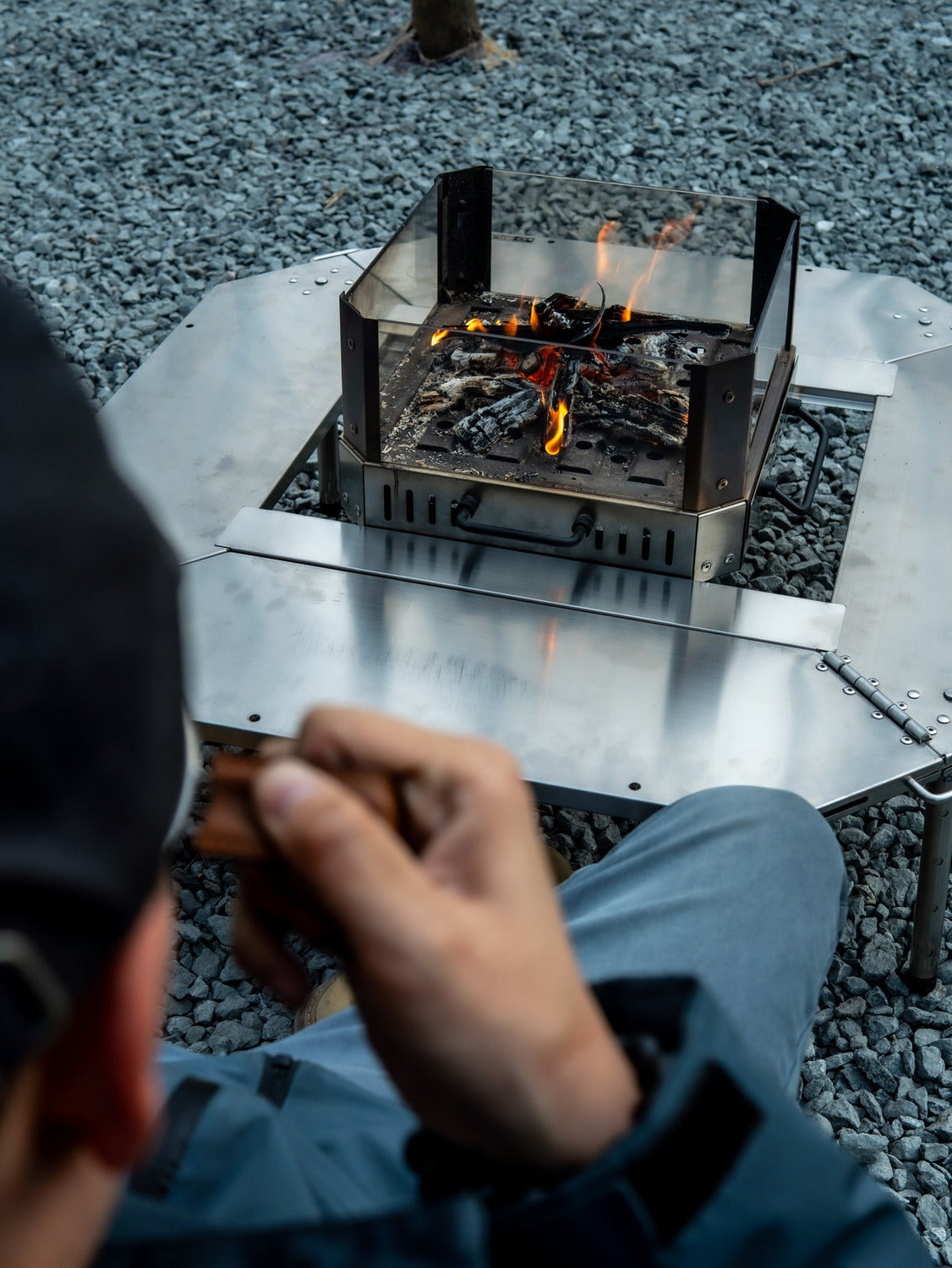 Seated view over the shoulder of someone feeding wood into a fire in the centre of the Biloc Octa table, with the inner table set fitted. The stove is a separate purchase.