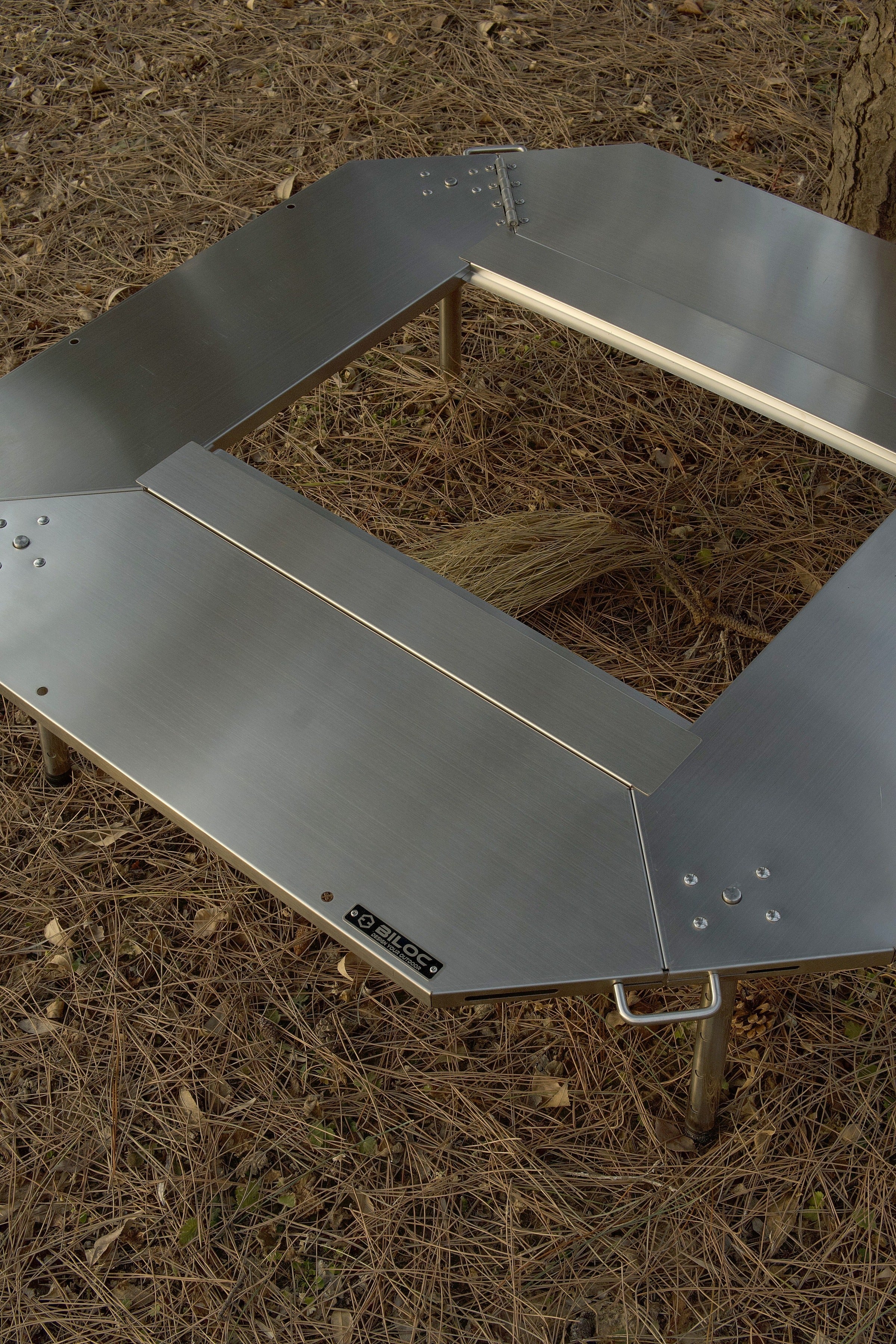 Octa table folded open and laid on pine-needle ground, seen from above, with the BILOC nameplate on the plate edge.