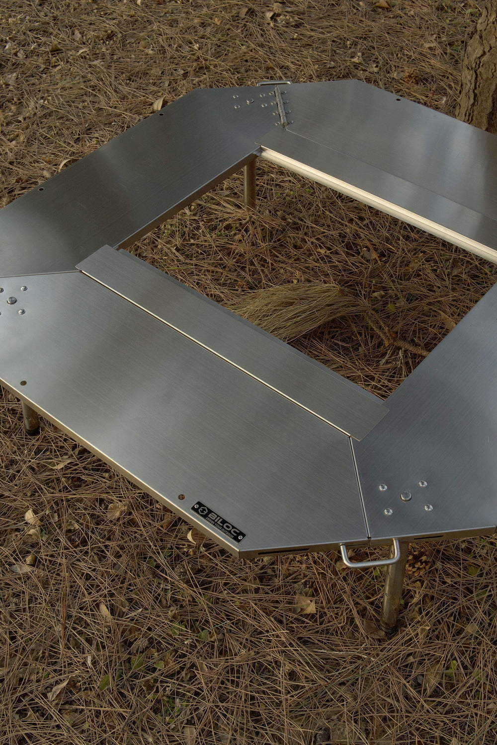 Octa table folded open and laid on pine-needle ground, seen from above, with the BILOC nameplate on the plate edge.