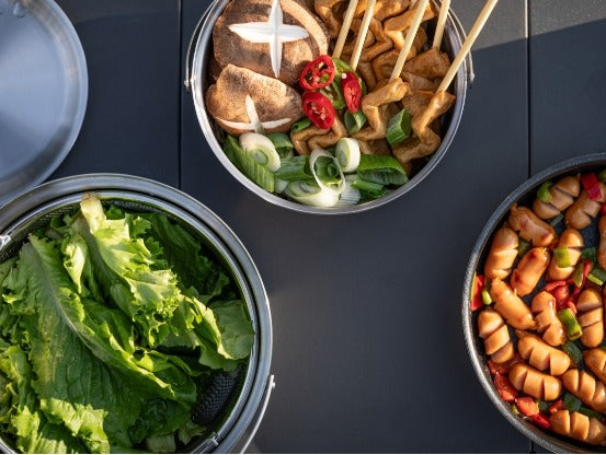 Top-down view of the cookware filled with fresh greens, skewers, and cooked sausages