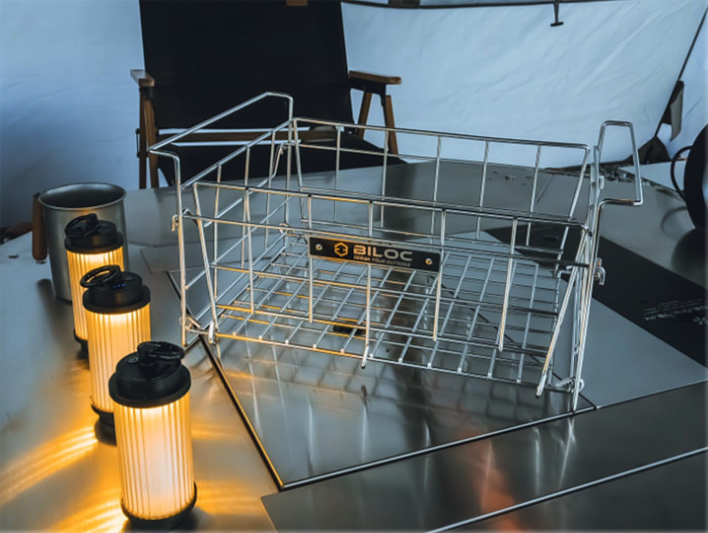 Folding wire basket standing on a camp table as an open bin, beside lanterns.
