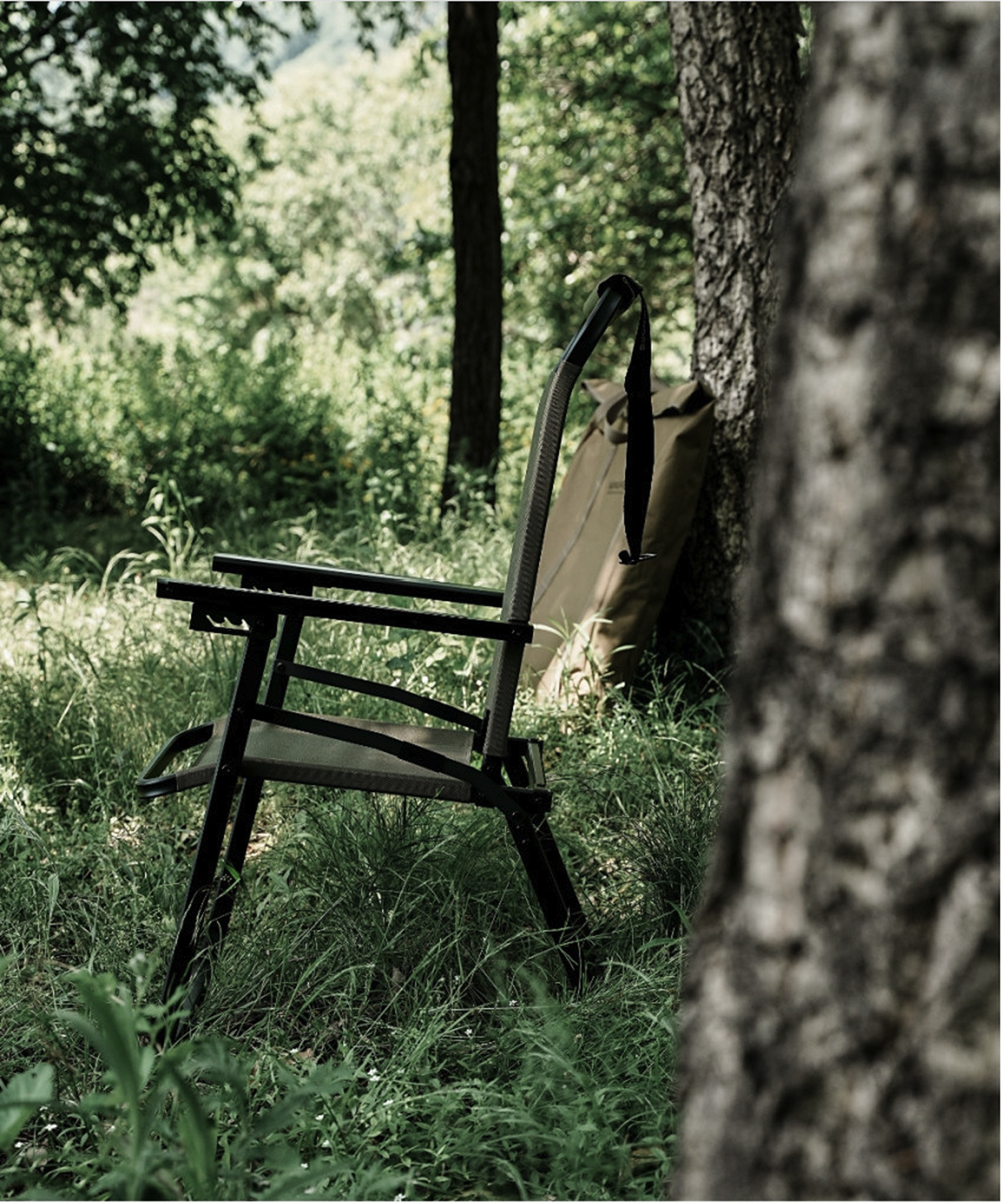 Black FLOW folding chair in a forest clearing with the roll-top storage bag against a tree