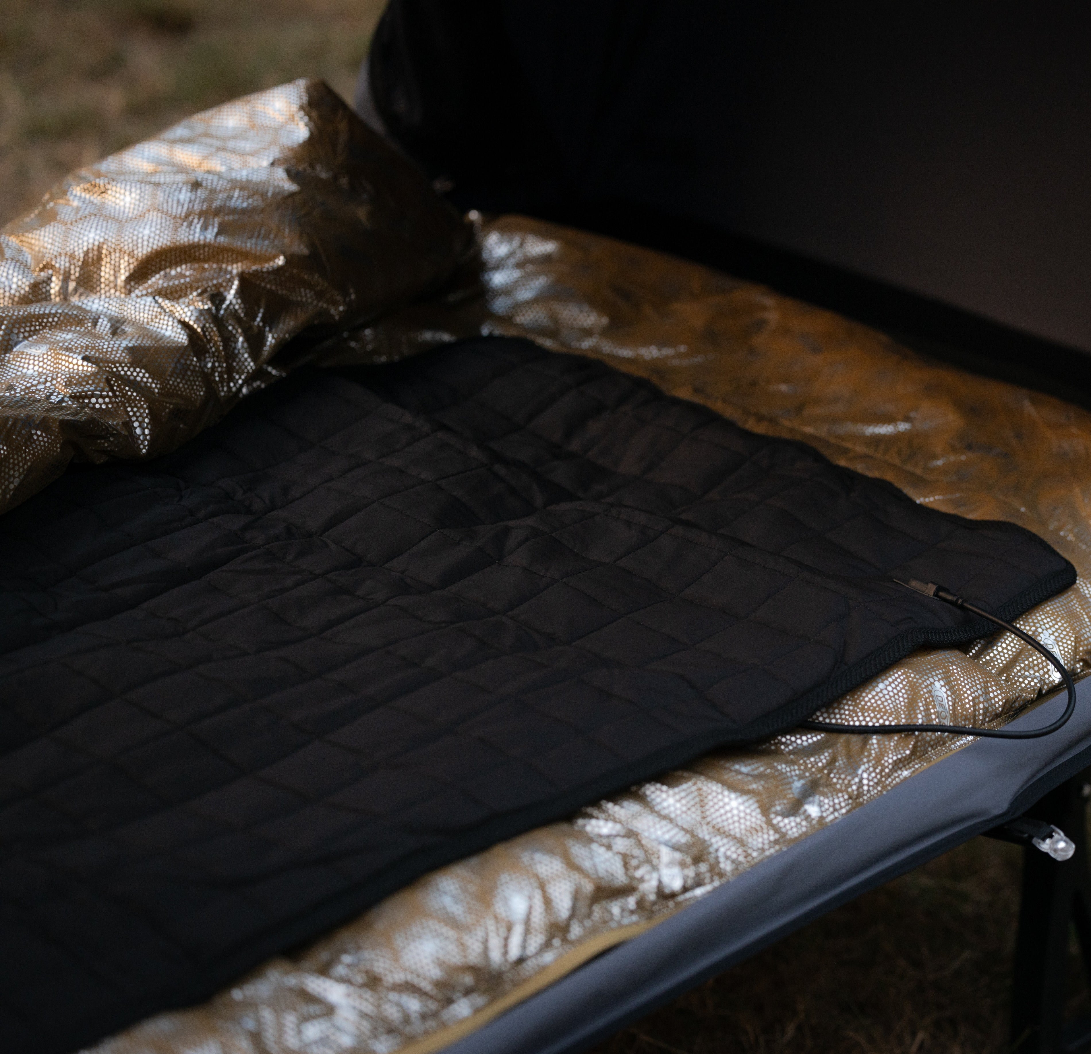The black quilted Carbon Heating Pad Large laid flat on a camp cot inside a shelter, its power cable running from the edge of the pad.