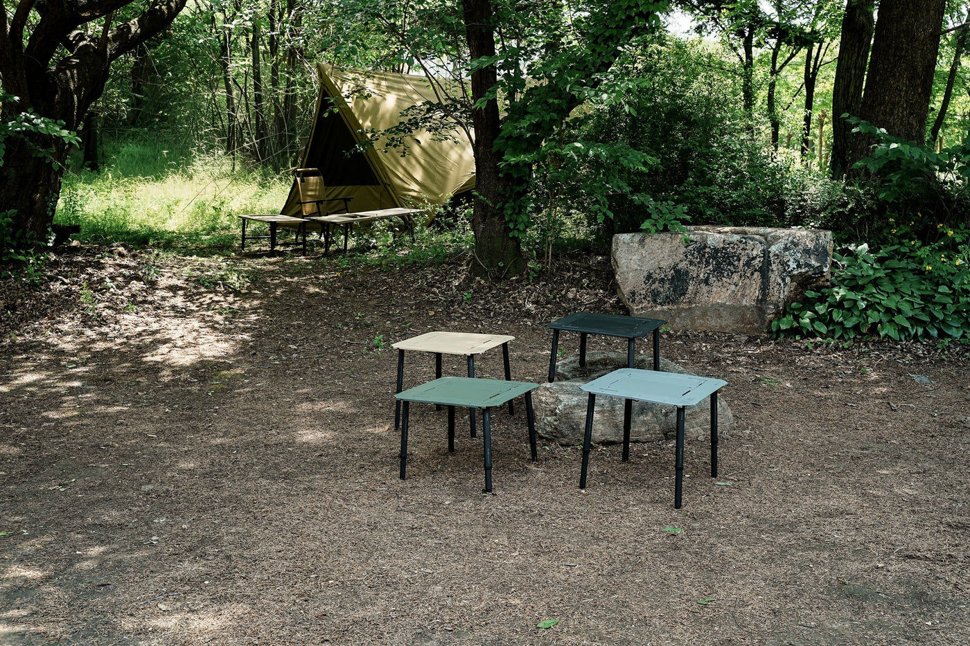 Four BUENOFE IGT Connect Table 1.5 standalone tables in all colorways grouped on the forest floor in front of a tan tent, a wide campsite scene showing the range set up in nature.