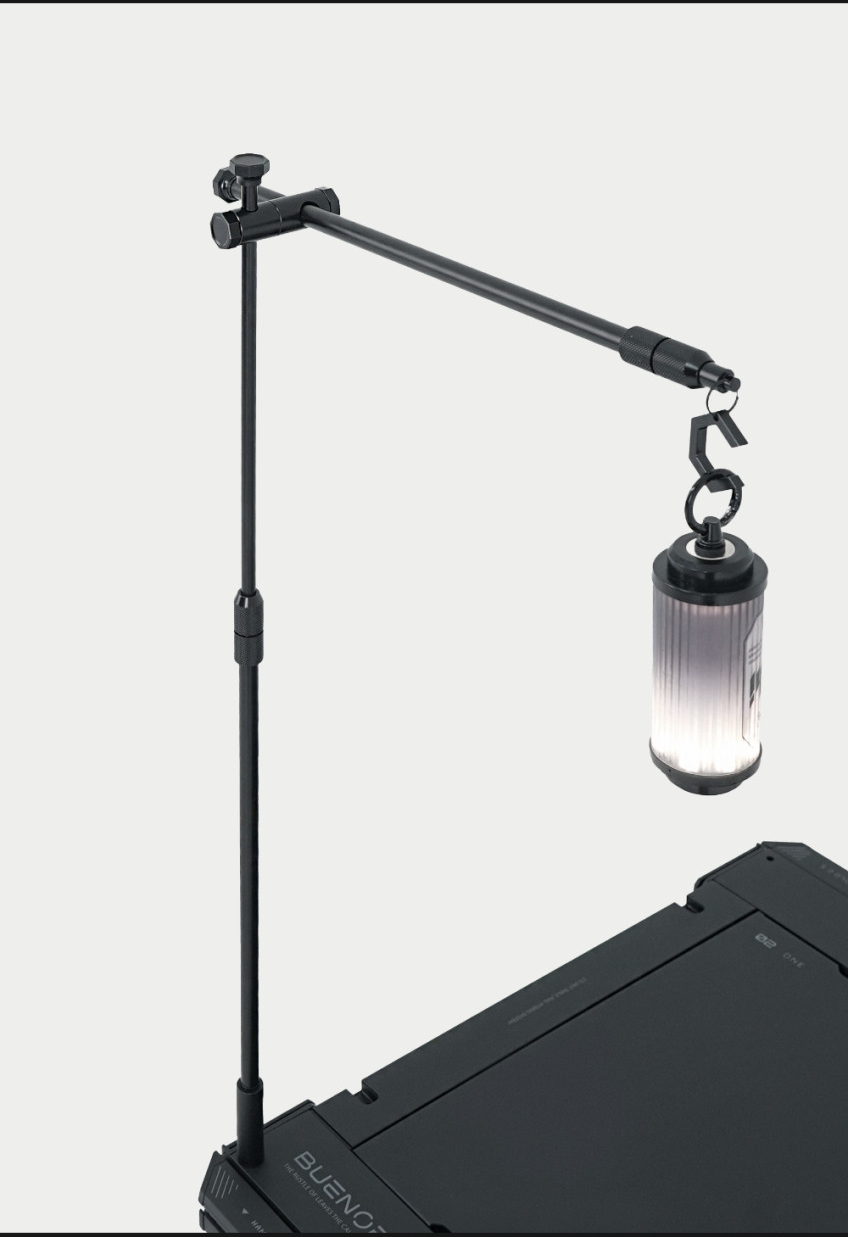 BUENOFE IGT Lantern Hanger shown close up with the boom arm and coupler holding a cylindrical lantern above a tilted dark table, a dramatic product-and-lantern styling shot on a white background.