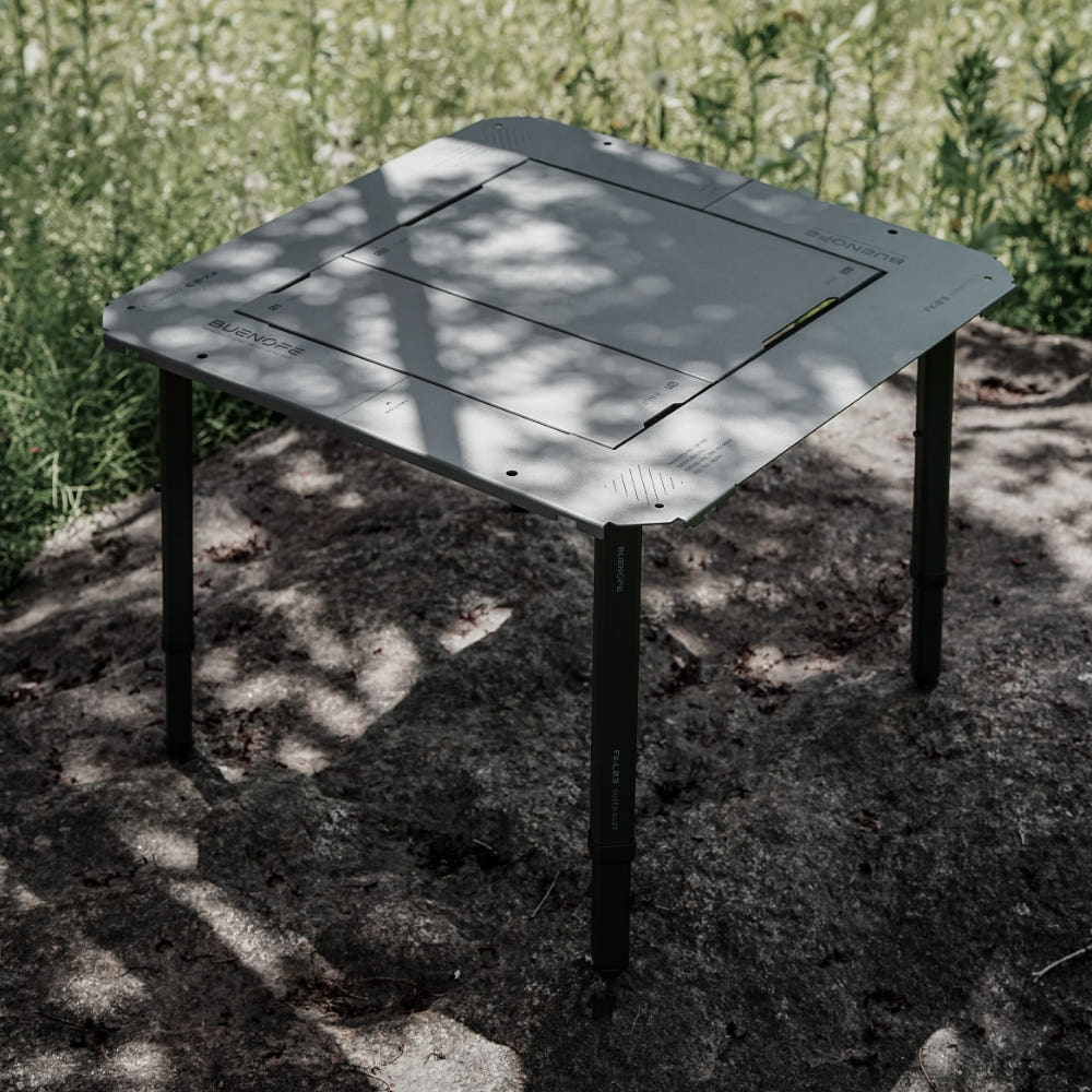 BUENOFE IGT Quick Slot Table Leg shown holding a gray IGT tabletop on a rocky, grassy patch in dappled sunlight, the black legs standing level on uneven natural ground outdoors.
