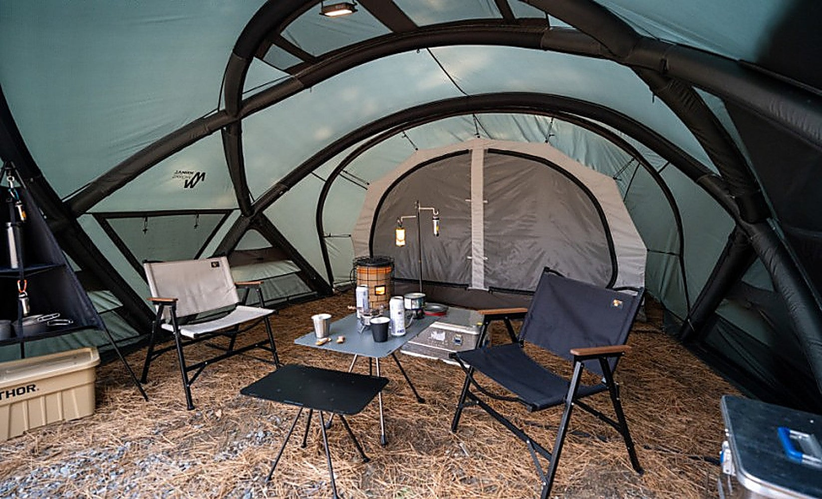 Inside the Shelter GM: black-sleeved frame arches under a sage canopy, inner tent, chairs and table on pine needles