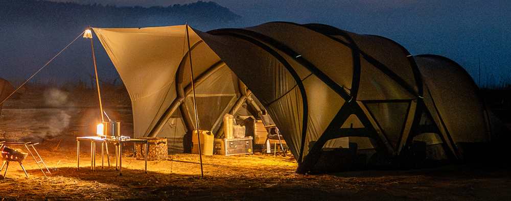 Wide illuminated shelter setup at night showing a large covered area with table and gear underneath