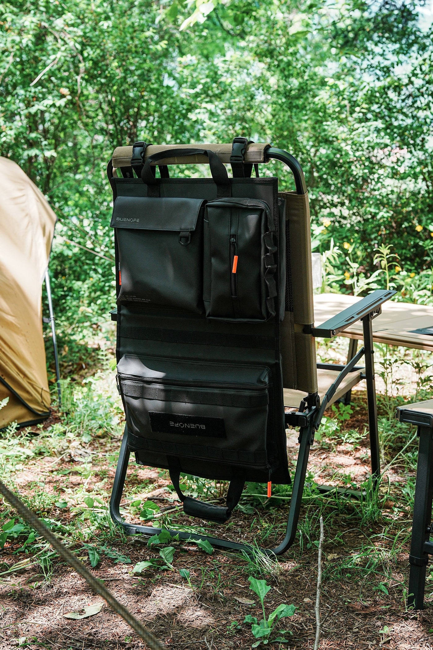 BUENOFE Multi Storage Bag Slim mounted on the back of a folding chair at a forest campsite, vertical view with a canvas tent to the left and a camp table to the right, showing the organizer in a real outdoor setting.