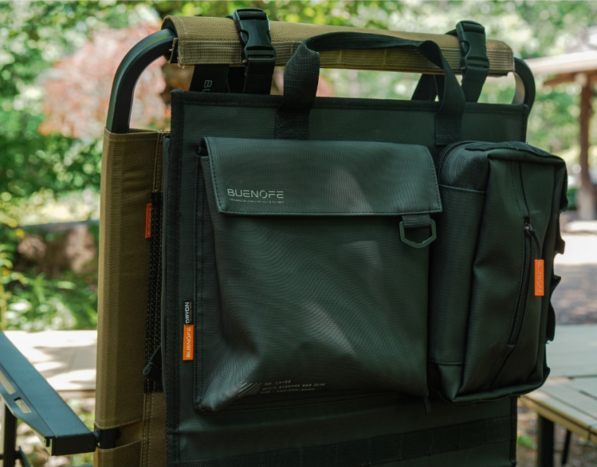 BUENOFE Multi Storage Bag Slim close-up while mounted on a camp chair back outdoors, showing the front flap pocket with the BUENOFE logo, the side tissue pouch and the orange zipper pulls in natural daylight.