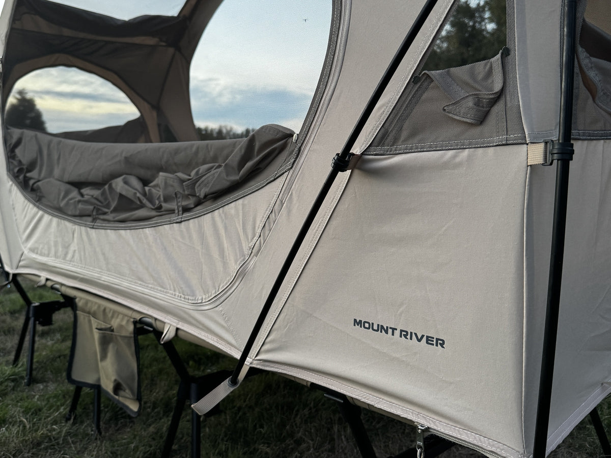 Side view of the Neo Nest 80 on a camp cot at dusk, oval window open and the Mount River logo on the panel (cot sold separately).