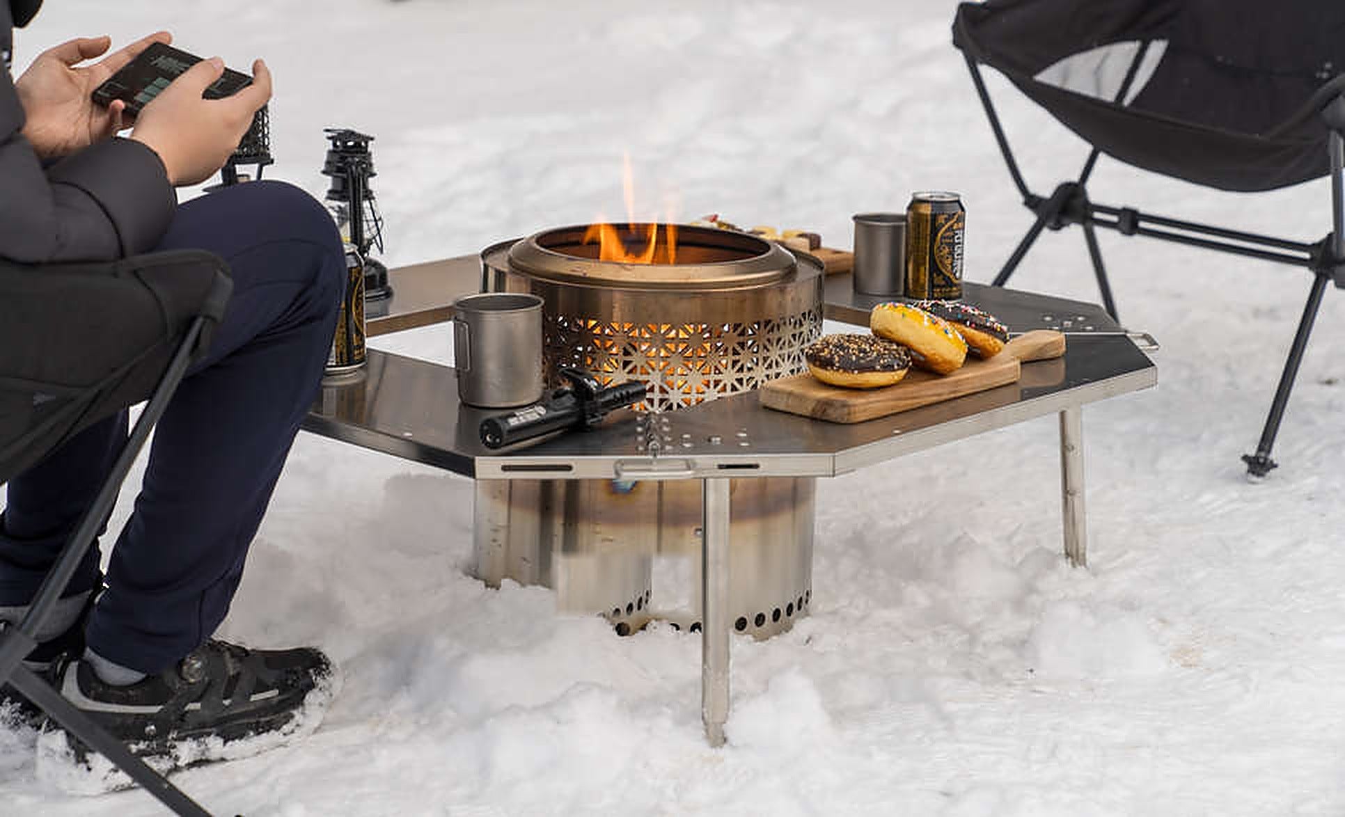 Sitting fireside at the Biloc Octa table in snow, with a wood stove burning in the centre and snacks on the plates. The stove and accessories shown are separate purchases.