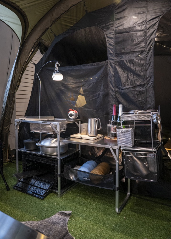 Stella shelf standing as a camp kitchen inside a shelter, loaded with cookware under a lamp arm. The gear shown is not included.