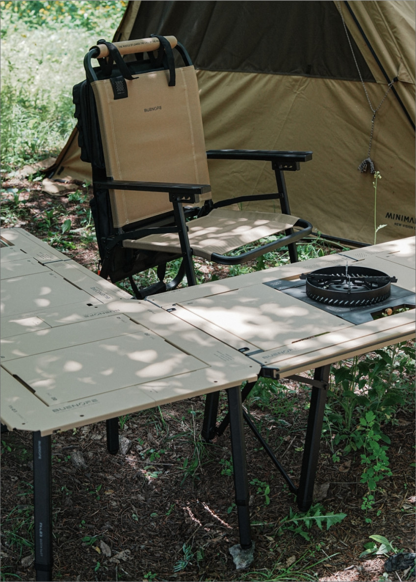 BUENOFE Stove Windscreen and BX-09 Table Burner set up on a tan IGT camp table beside a folding camp chair and tent in a sunlit forest clearing, showing the cooking system in a real outdoor campsite.